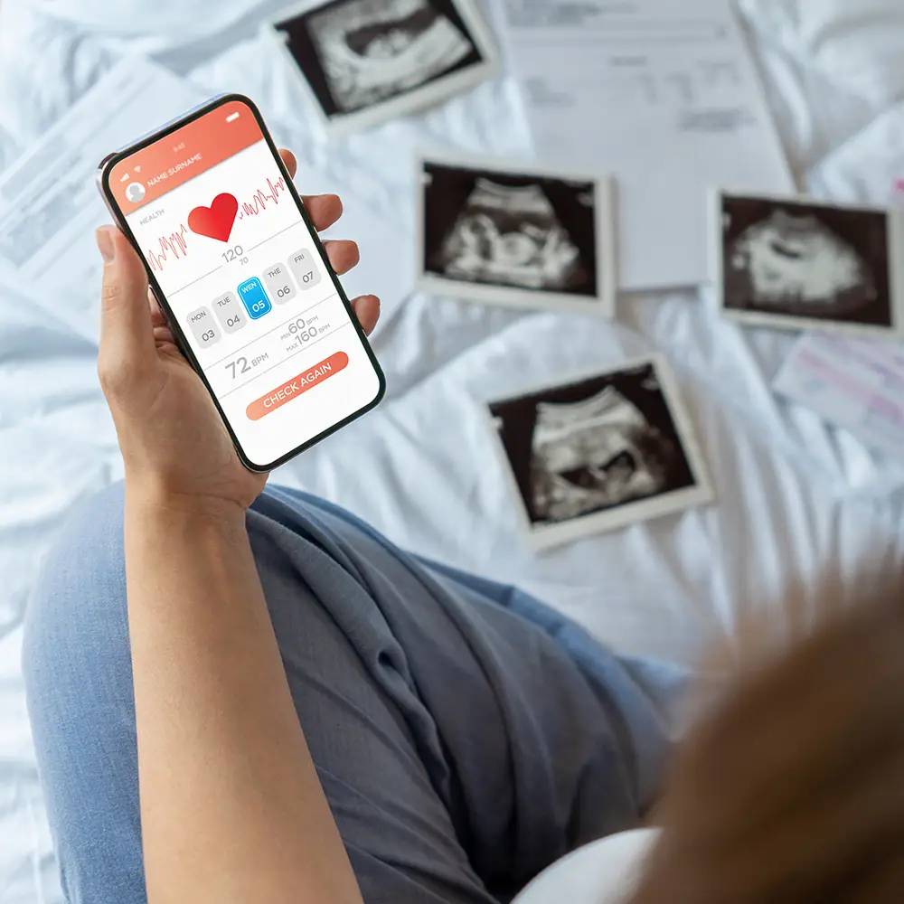 Pregnant woman holding a smartphone with a healthcare app, surrounded by ultrasound images on a bed.